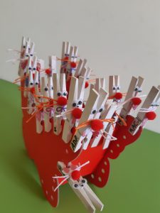 Zdjęcie przedstawia króliczki zrobione z białych klamerek w czerwonym tekturowym wózku na biało zielonym tle.