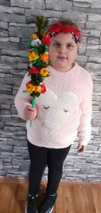 Na tle kamiennej ściany stoi uczennica trzymająca w prawej dłoni wykonaną przez siebie palmę wielkanocną.