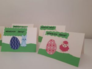 Na stoliku ustawiono cztery kartki świąteczne. Na dwóch z pierwszego planu widać napis na zielonym tle "Wesołych Świąt" , naklejone pisanki oraz wielkanocne zwierzątka.