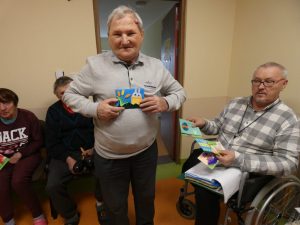 Starszy Pan stoi na środku sali i trzyma przed sobą otrzymaną od uczniów kartkę. Obok na wózku siedzi Pan przeglądający różne kartki świąteczne.