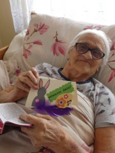 Na łóżku leży starsza Pani trzymająca w dłoni otrzymaną kartkę świąteczną.