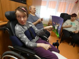 Na pierwszzym planie Pani na wózku trzyma wykonaną przez uczniów kartkę. W tl;e starsza Pani także trzyma kartkę świąteczną.