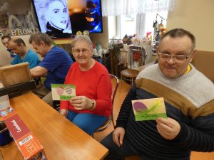 Przy stole siedzą seniorzy. Dwójka z nich trzyma w ręku kartki zrobione przez uczniów.