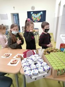 Uczniowie stoją w kolejce obok stolików na których umieszczono karton z szoczkami, paletę jogurtów oraz tosty.