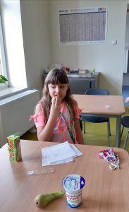 Na zdjęciu uczennica sziedzi za stołem i je tosta. Na stole leżą gruszka, jogurt oraz soczek w kartoniku.