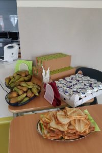 Zdjęcie dwóch szkolnych stolików, na których umieszczono talerz z tostami, podłużne miski z gruszkami, paletę jogurtów oraz kartony z soczkami.