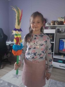 Uczennica stoi w swoim pokoju i trzyma w prawej dłoni wykonaną przez siebie palmę wielkanocną.