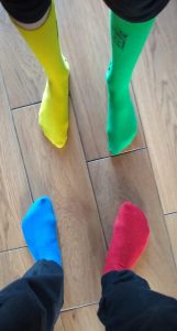 Na drewnianej podłodze stoja cztery stopy - każda w innej skarpetce: niebieskiej, różowej, zielonej i żółtej.