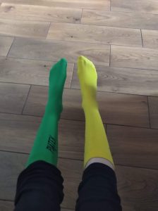 Na tle drewnianej podłogi dwie nogi ubrane w dwie różne podkolanówki - zieloną i żółtą.