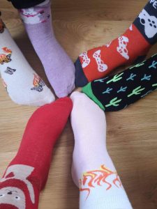 Na drewnianej podłodze ustawione są stopy trzech osób stykające sie palcami i ubrane w różnokolorowe skarpetki.