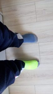 Na tle drewnianej podłogi widoczne są dwie nogi ze stopami w różnych skarpetkach - granatowej i zielonej.