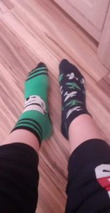 Na tle drewnianej podłogi widoczne dwie nogi w dwóch róznych skarpetkach - zielonej i granatowej z wzrorkami.