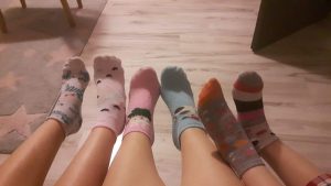 Na tle drewnianej podłogi widoczne są uniesione nogi trzech osób ubrane w kolorowe skarpetki.