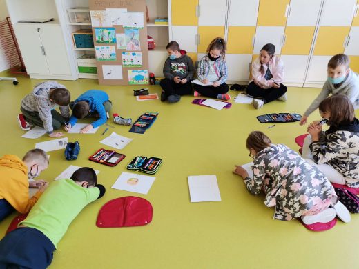 Zdjęcie przedstawia uczniów siedzących lub leżących na podłodze sali lekcyjnej. Przed nimi leżą kartki i piórniki.