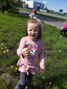 Uczennica stoi z otwartą buzią na trawniku. W wyciagniętej prawej dłoni trzyma dmuchawca. W tle widać budynek szkoły.