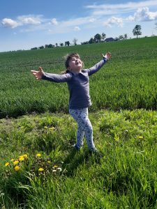 Uczennica z rozłożonymi rękami i twarzą skierowaną ku słońcu stoi na łące.