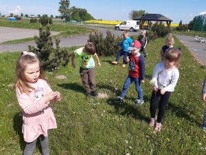 Dzieci zbierają dmuchawce. Stoją na wąskim pasie zieleni z obu stron ograniczonego chodnikami.