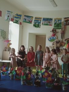 Uczniowie stoją w rządku. Za nimi ściana ich klasy ozdobiona dużymi kukiełkami oraz pracami uczniów, a przed nimi stół na którym umieszczono własnoręcznie wykonane przez nich prezenty dla mam.