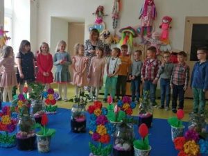Uczniowie stoją w rządku. Za nimi ściana ich klasy ozdobiona dużymi kukiełkami, a przed nimi stół na którym umieszczono własnoręcznie wykonane przez nich prezenty dla mam.