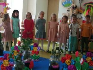 Uczniowie stoją w rządku. Za nimi ściana ich klasy ozdobiona dużymi kukiełkami, a przed nimi stół na którym umieszczono własnoręcznie wykonane przez nich prezenty dla mam.