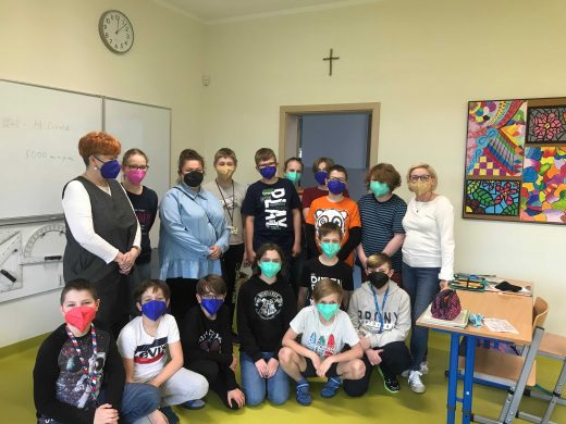 Zdjecie grupowe - uczniowie w otrzymanych maseczkach, ich nauczycielka, Pani Dyrektor i przedstawicielka firmy Sohbi Craft Poland, pozują wspólnie do zdjęcia w sali lekcyjnej.