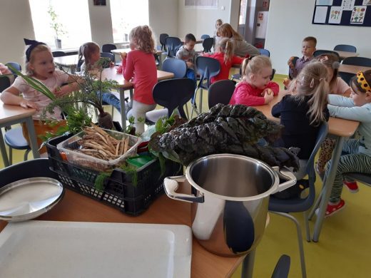 Uczniowie z oddziału przedszkolnego siedzą przy stolikach w szkolnej stołówce. Na pierwszym planie widzimy stół z ustawionym na nim stalowym garnkiem oraz plastikową skrzynkę z warzywami.