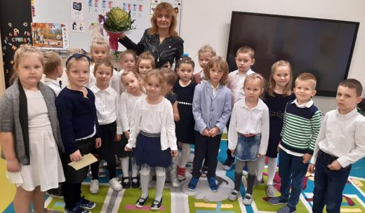 Wychowawczyni oddziału przedszkolnego trzyma w wysoko podniesionej dłoni bukiet otrzymany od uczniów. W okół niej stoją jej wychowankowie i wspólnie pozują do zdjęcia.