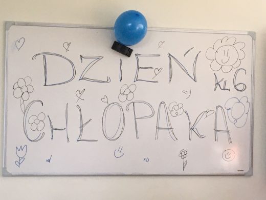 Zdjęcie białej tablicy z odręcznym napisem "DZIEŃ CHŁOPAKA KL 6". U góry tablicy przyczepiono za pomocą gąbki niebieski balonik.