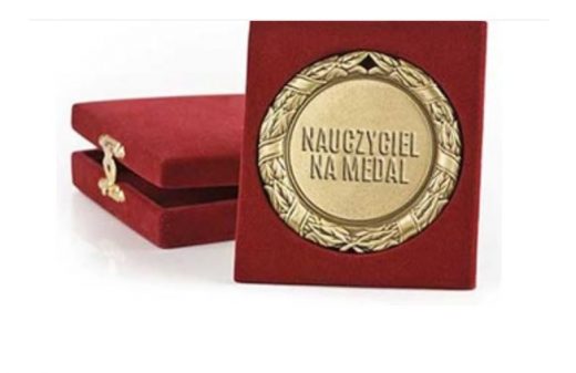 Grafika przedstawia czerwone aksamitne pudełko zamykane na srebrną zasuwkę oraz oparty o nie medal z napisem NAUCZYCIEL NA MEDAL, umieszczony w czerwonej, aksamitnej ramce.