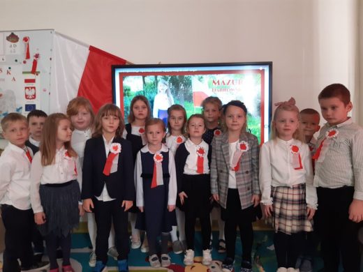 Uczniowie pozują do zdjęcia w strojach galowych z przyczepionymi do piersi kotylionami. WE tle widać tablicę interaktywną.