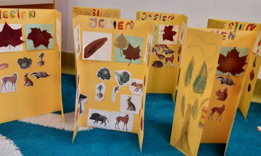 Zdjęcie lapbooków wykonanych przez uczniów oddziału przedszkolnego. Prace zostały ustawione obok siebie w pozycji pionowej. Każda z nich została wykonana z żółtej kartki papieru i ozdobiona odręcznymi rysunkami.