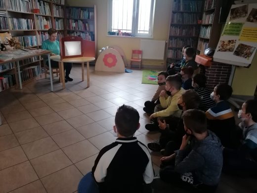 Zdjęcie wykonane w Gminnej Bibliotece Publicznej. Pani Bibliotekarka siedzi przy stoliku na którym ustawiono biały ekran. Uczniowie siedzą na podłodze na przeciwko.