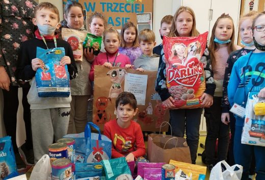 Uczniowie pozują do zdjęcia w jednej z klas. Przed nimi na podłodze ustawiono zebraną karmę. Dzieci trzymają w dłoniach przysmaki dla zwierząt oraz plakat przedstawiający pieski.