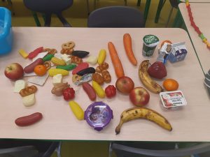Na stoliku leżą owoce, warzywa, kefir, mleko i różne serki oraz plastikowe rybki i precelki. Wszystkie te przedmioty dzieci ułożą w stworzonej przez siebie piramidzie żywienia.