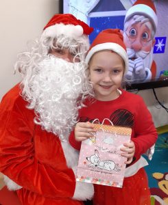 Uczennica oddziału przedszkolnego w czerwonej czapeczce i otrzymaną torebką z prezentami pozuje do zdjęcia z Mikołajem.