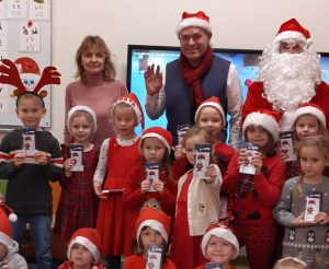 Uczniowie oddziału przedszkolnego wraz z wychowawczynią, Świętym Mikołajem i jego pomocnikiem pozują do zdjęcia. Dzieci mają na głowach czerwone czapeczki a w dłoniach trzymają czekolady.