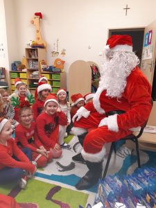 Święty Mikołaj siedzi na krzesełu a przed nim na dywanie siedzą uczniowie oddziału przedszkolnego w czerwonych czapeczkach.