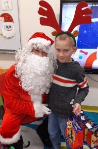 Uczeń oddziału przedszkolnego w opasce z przyczepionymi rogami renifera i otrzymaną torebką z prezentami pozuje do zdjęcia z Mikołajem.