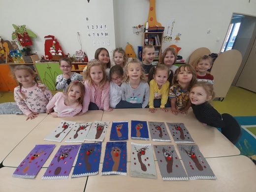 Uczniowie oddziału przedszkolnego stoją w grupie i pozują do zdjęcia. Przed nimi na złączonych stolikach leżą ułożone w dwóch rzędach ich prace przedstawiające renifery.