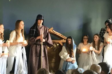 Zdjęcie wykonane podczas szkolnych Jasełek na sali teatralnej. Na tle stajenki stoi uczeń Przebrany za Józefa, obok siedzi Maryja z dzieciątkiem a wokół nich stoją uczennice ubrane na biało i trzymające w dłoniach zapalone świece.
