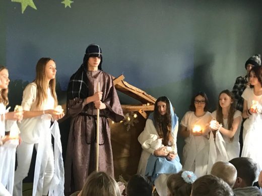 Zdjęcie wykonane podczas szkolnych Jasełek na sali teatralnej. Na tle stajenki stoi uczeń Przebrany za Józefa, obok siedzi Maryja z dzieciątkiem a wokół nich stoją uczennice ubrane na biało i trzymające w dłoniach zapalone świece.