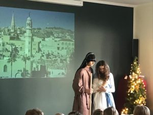 Zdjęcie wykonane podczas szkolnych Jasełek na sali teatralnej. Dwoje uczniów przebranych za Józefa i Maryję stoi na tle czarnej ściany na której wyświetlono zdjęcie starożytnego miasta.