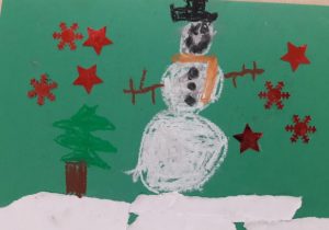 Świąteczna kartka wykonana przez ucznia oddziału przedszkolnego. Na zielonym tle czerwone gwiazdki oraz biały bałwan i zielona choinka.
