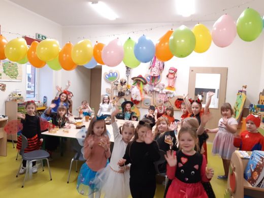 Dzieci z oddziału przedszkolnego rozproszone po sali machaja do zdjęcia. Nad nimi wisi łańcuch utworzony z kolorowych balonów.