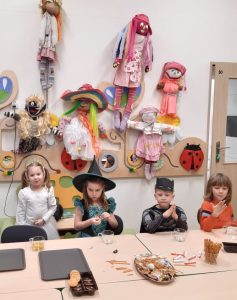 Dzieci z oddziału przedszkolnego siedzą za stołem na którym ustawiono przekąski. W tle widać ścianę na której umieszczono kolorowe lalki.