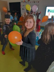 Dzieci z oddziału przedszkolnego bawią się kolorowymi balonami. W centralnym punckie stoi uczennica patrząca się w stronę aparatu i trzymająca w dłoniach pomarańczowy balonik.