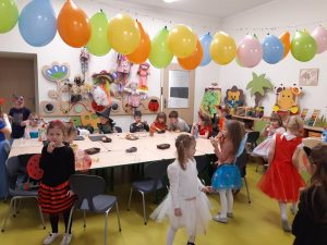 Dzieci z oddziału przedszkolnego bawią się przy stole z przekąskami. Nad stołem zawieszono na sznurku kolorowe balony.