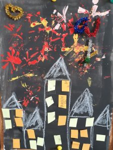 Zdjęcie pracy ucznia oddziału przedszkolnego- na czarnym tle namalowane biało kretką wysokie domy a nad nimi kolorowe fajerwerki.