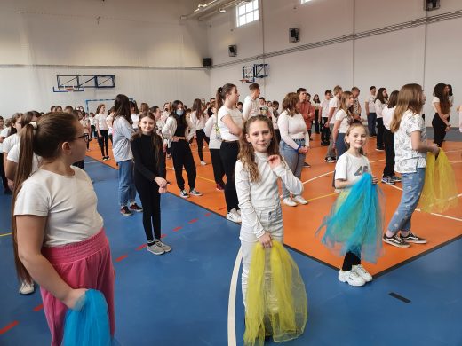 Uczniowie w rzędach stoją na sali gimnastycznej. Dziewczynki stojące na pierwszych miejscach trzymają żółto- niebieskie tiulowe kawałki materiału.