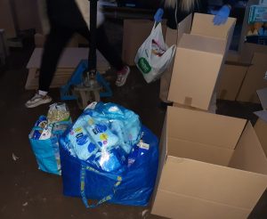 Pakowanie darów dla mieszkańców Ukrainy. Na zdjęciu kartony oraz stojące przy nich dwie torby wypełnione darami. W tle postać, która w jednej dłoni trzyma plastikową torbę a w drugiej karton.